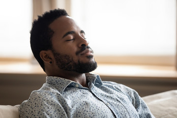 Tranquil young african man resting eyes closed breathing on couch - obrazy, fototapety, plakaty