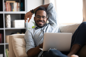 Smiling young adult african guy using laptop relaxing on sofa