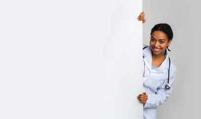 Young female medical intern looking out of behing white advertising board