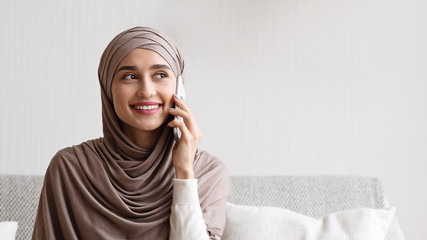 Young muslim woman in headscarf having pleasant phone conversation at home