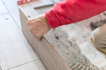 Man installing rectangular shaped floor tiles in kitchen. Applying adhesive before installation and verifying afterwards