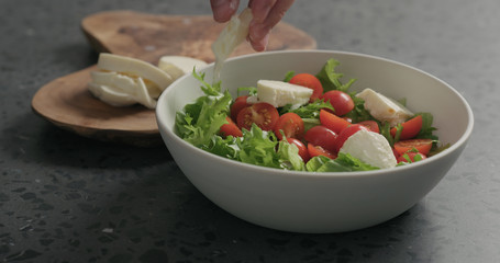 man hands preparing salad with mozzarella, cherry tomatoes and frisee leaves in white bowl on terrazzo surface