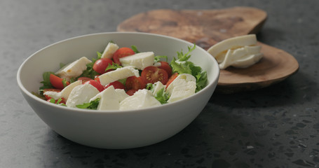 preparing salad with mozzarella, cherry tomatoes and frisee leaves in white bowl on terrazzo surface