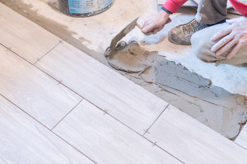 Man installing rectangular shaped floor tiles in kitchen. Applying adhesive before installation and verifying afterwards