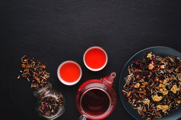 Aromatic fresh brew natural red fruit tea in two bowls ,glass teapot,glass jar and plate with dry tea leaves on a black concrete background