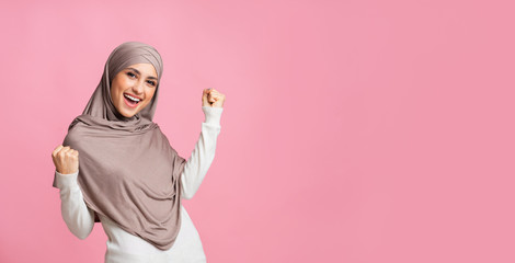 Muslim girl in headscarf celebrating success, raising hands and shaking fists