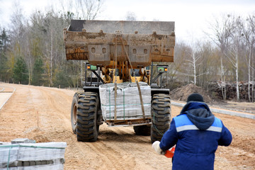 Fototapeta premium The tractor lifted the bucket tray with paving slabs, the worker monitors the progress of work.