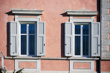 Two italian windows on the pink color wall facade with open wooden white shutters