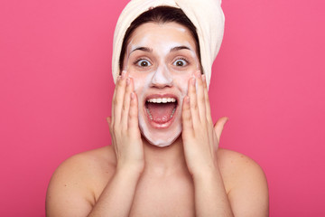 Studio shot of pretty girl with facial mask, surprised female with white towel on her head, lady having astonished expression, posing isolated over pink background. Skin care and beauty concept.