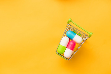 Small shopping cart full of pills on yellow background. Medicine and pharmacy concept