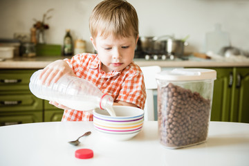 Little boy sitting in the kitchen, having breakfast, pouring milk into a cereal cup, preparing...
