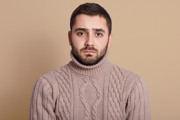 Horizontal shot of attractive young man with beard posing in studio against beige wall, looking at camera with serious facial expression, wearing knitted sweater, being in bad mood, having problems.