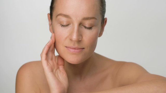 Close-up beauty portrait of young woman with smooth healthy skin, she gently touches her face with her fingers on white background and smiles