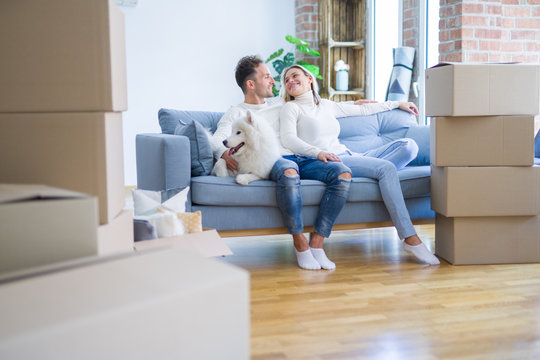 Young beautiful couple with dog sitting on the sofa at new home around cardboard boxes