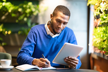 Black man holding tablet and writing in notebook
