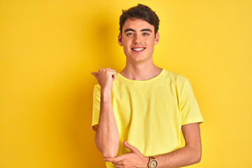 Teenager boy wearing yellow t-shirt over isolated background smiling with happy face looking and pointing to the side with thumb up. © Krakenimages.com