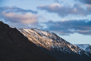 Fototapeta premium Atmospheric alpine landscape to snowy mountain ridge in sunset. Snow shines in golden light on mountain peak. Beautiful shiny snowy top. Evening cloudy sky. Wonderful scenery in gold sunset shades.