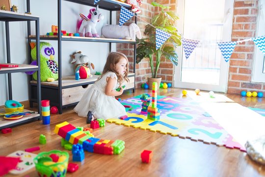 Adorable blonde toddler playing with building blocks toy around lots of toys at kindergarten