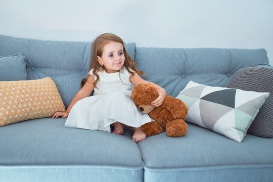 Adorable blonde toddler sitting on the sofa playing with teddy bear at home