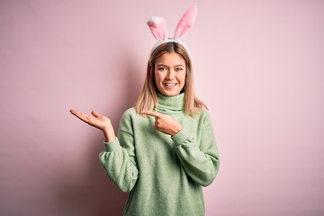 Young beautiful woman wearing easter rabbit ears standing over isolated pink background amazed and smiling to the camera while presenting with hand and pointing with finger.