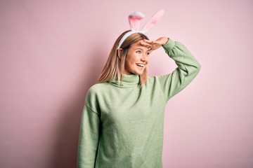 Young beautiful woman wearing easter rabbit ears standing over isolated pink background very happy and smiling looking far away with hand over head. Searching concept.
