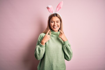 Young beautiful woman wearing easter rabbit ears standing over isolated pink background smiling...