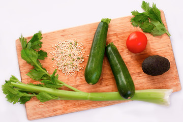 Green vegetables (celery, cucumbers, zucchini, avocado) and a tomato on a wooden chopping board