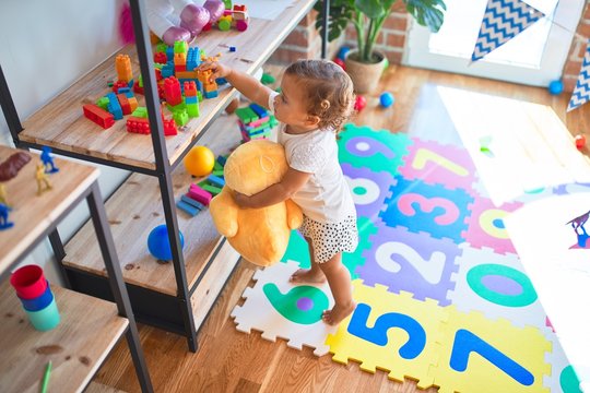 Adorable toddler holding duck doll standing around lots of toys at kindergarten