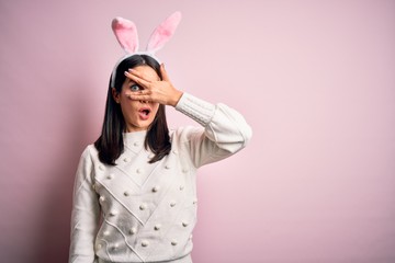 Young caucasian woman wearing cute easter rabbit ears over pink isolated background peeking in shock covering face and eyes with hand, looking through fingers with embarrassed expression.