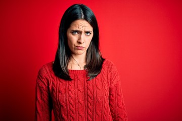 Young brunette woman with blue eyes wearing casual sweater over isolated red background skeptic and...