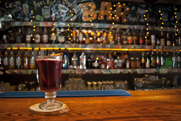 Red cocktail on bar counter. Mulled wine with fruits and cinnamon on the background of bottles with alcohol. Glass with booze on a wooden board. Cinnamon burns and smokes.