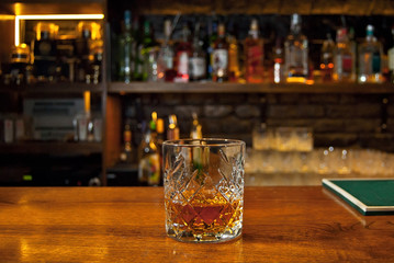 Yellow cocktail on bar counter. Whiskey, rum or brandy on the background of bottles with alcohol. Glass with booze on a wooden board.