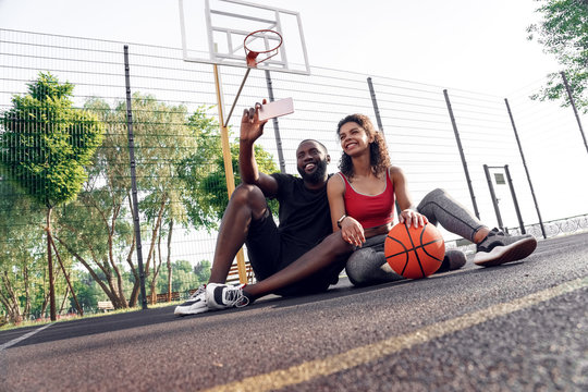 Outdoors Activity. African couple sitting on basketball court taking selfie on smartphone smiling happy