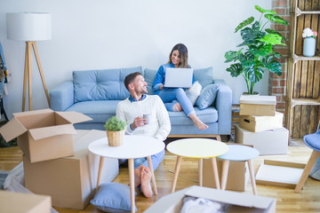 Young beautiful couple sitting on the sofa drinking cup of coffee using laptop at new home around cardboard boxes