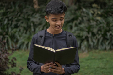 A young boy is reading a black notebook in a park over blurred background