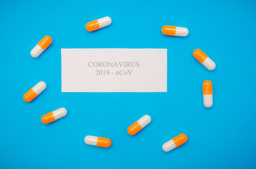 white sheet of paper with the inscription Coronavirus among orange-and-white capsules on a blue background. Medical background, top view