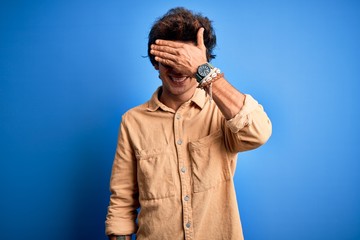 Young handsome man wearing casual shirt standing over isolated blue background smiling and laughing with hand on face covering eyes for surprise. Blind concept.