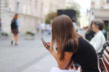 A young girl is looking into a smartphone that is holding. A woman sits on a bench in the center of an ancient city against a motorcycle and passers-by in a blurred blur. Lifestyle and leisure