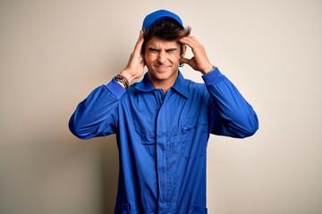 Young mechanic man wearing blue cap and uniform standing over isolated white background suffering from headache desperate and stressed because pain and migraine. Hands on head.