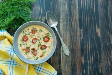 Cheese soup with fried sausages and herbs. Tasty cream cheese soup in a white plate. Dinner table. The balance of healthy eating. Copy space. Wood background.