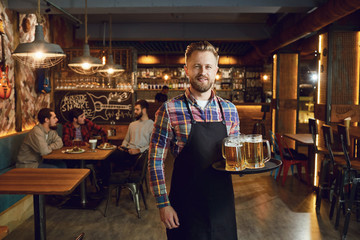 Bearded waiter with a tray of glasses of beer against the background of a pub bar - obrazy, fototapety, plakaty