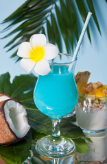 Exotic tropical Blue Curacao cocktail drink in a glass with Plumeria frangipani flower, palm leaf, fresh coconut on the background.