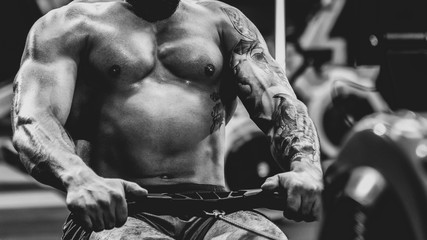 Fototapeta na wymiar close up Young muscular man with tattoo doing exercises on rowing machine in gym, dramatic black and white image