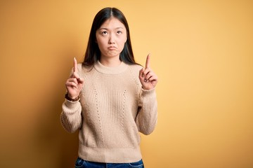 Young beautiful asian woman wearing casual sweater over yellow isolated background Pointing up looking sad and upset, indicating direction with fingers, unhappy and depressed.