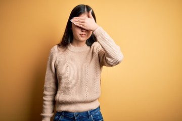 Young beautiful asian woman wearing casual sweater over yellow isolated background covering eyes with hand, looking serious and sad. Sightless, hiding and rejection concept