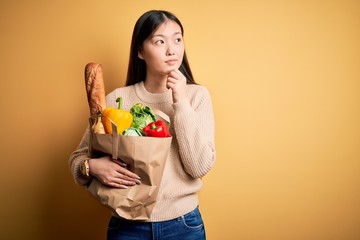 Young asian woman holding paper bag of fresh healthy groceries over yellow isolated background with hand on chin thinking about question, pensive expression. Smiling with thoughtful face. Doubt 