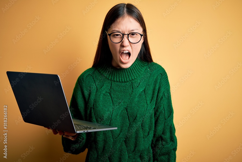 Poster Young asian business woman wearing glasses and working using computer laptop angry and mad screaming frustrated and furious, shouting with anger. Rage and aggressive concept.