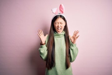 Young asian woman wearing cute easter bunny ears over pink background celebrating mad and crazy for success with arms raised and closed eyes screaming excited. Winner concept