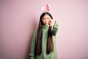 Young asian woman wearing cute easter bunny ears over pink background looking unhappy and angry showing rejection and negative with thumbs down gesture. Bad expression.