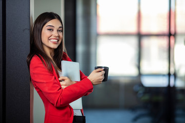 Beautiful business woman in the coffee break. Young business woman enjoying cup of coffee in the office. Beautiful young business woman smiling and holding laptop and coffee cup.